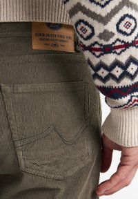 Pantalones de pana color caqui con una superficie texturizada, un parche de marca de cuero y un bolsillo trasero con detalles de costura tonal.