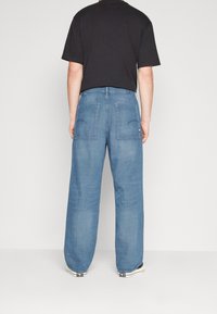 Person som bär löst sittande blå denimjeans med stora bakfickor och en svart t-shirt, stående mot en enkel vit bakgrund.