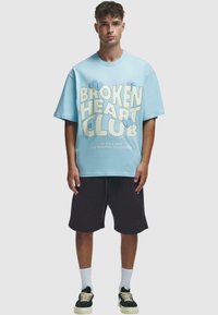 Šviesiai mėlyna oversized marškinėliai su grafika "BROKEN HEART CLUB", juodos šortai ir juodi sportbačiai, su baltais kojiniais ir laisvu stiliumi.