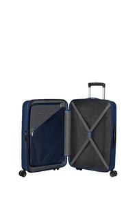 Valigia blu navy con un esterno rigido, scomparti con cerniera, cinghie interne e tasca in rete; dotata di quattro ruote e manico retrattile.