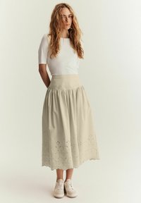 Beige midi kjol med en utsvängd design och broderat blommönster i nederkanten, ihopparat med en åtsittande vit topp och vita sneakers.