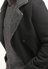 Svart ullrock med en strukturerad, fuzzy grå krage. Rocken har en dubbelknäppt design och stora knappar, i kombination med en stickad grå tröja.