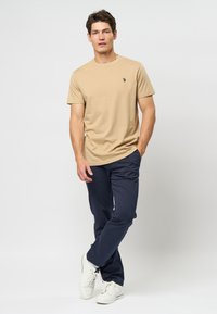 Beige crew neck t-shirt med lille sort logo, parret med mørkeblå bukser og hvide sneakers. Enkel design, blødt stof, afslappet udseende.