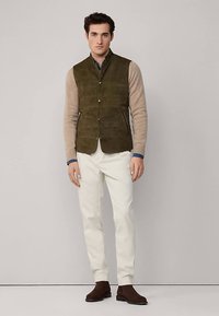 Veste en daim vert à boutons sur le devant, associée à un pull beige et un pantalon blanc. Des bottines marron complètent la tenue.