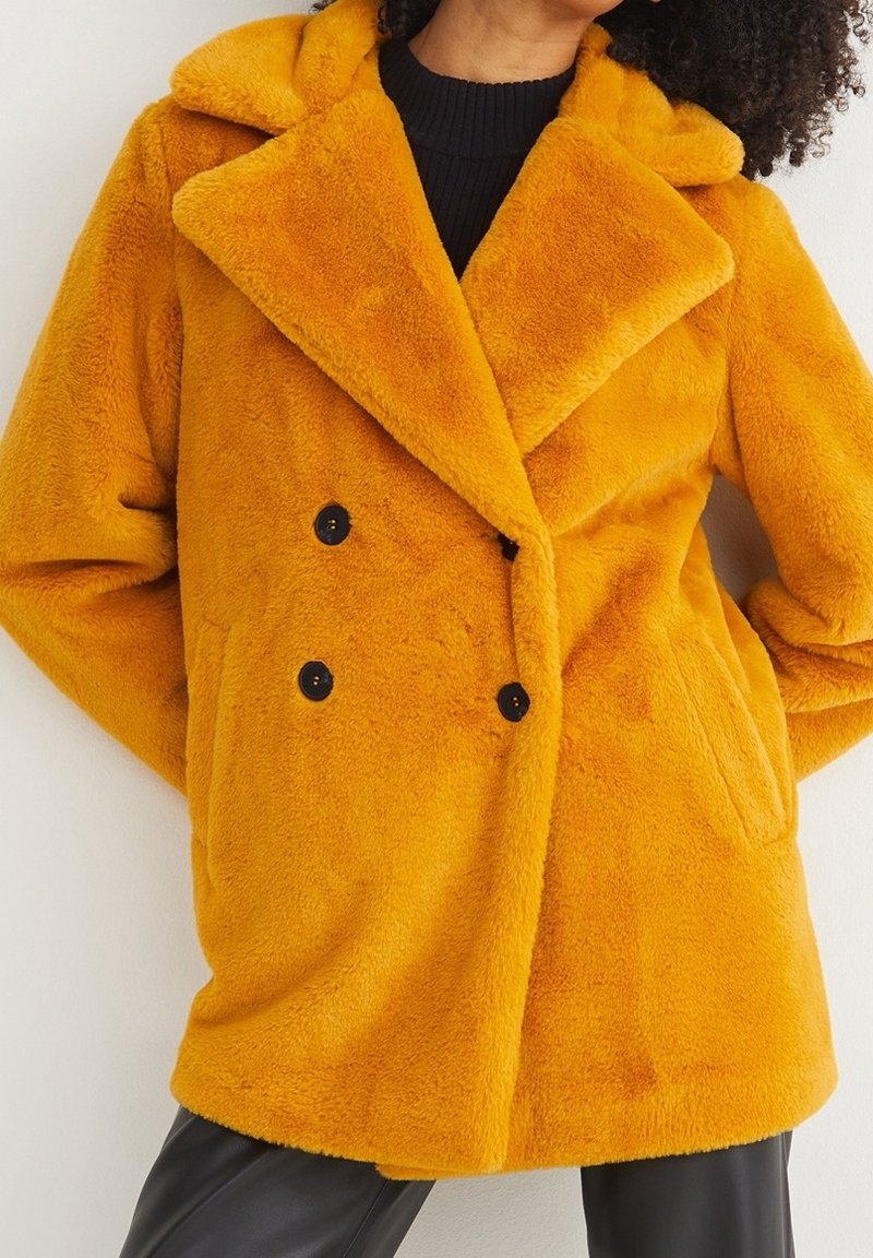 Personne portant un manteau en fausse fourrure orange vif à double boutonnage avec des boutons noirs par-dessus un haut noir.