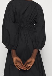 Svart klänning med figurnära midja, puffärmar och rynkade detaljer. Slät tyg med matt finish. Händerna placerade bakom ryggen.