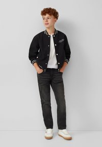 Giacca varsity nera con accenti a strisce bianche, abbinata a una t-shirt bianca e jeans scuri. Sneakers bianche completano l'outfit.