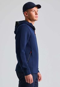 Felpa blu navy realizzata in tessuto morbido, dotata di chiusura con zip, due tasche laterali con dettagli rossi e cappuccio. Texture morbida, design casual.