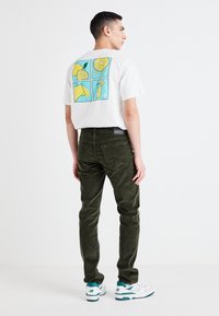 Balta T-kreksa ar krāsainu augļu grafiku aizmugurē; tumši zaļi korduroja bikses; baltie un tirkīza sporta apavi.