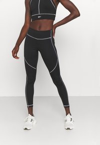Czarny sportowy crop top z szarymi przeszyciami, zestawiony z czarnymi legginsami o wysokim stanem z akcentowanymi szwami. Model nosi białe sneakersy.