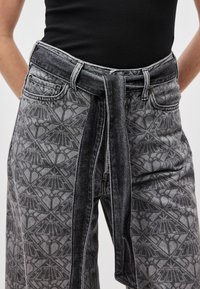 Gray patterned denim pants with a wide belt, featuring a floral design. Textured fabric with a relaxed fit and side pockets.