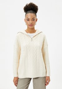 Crème kabelgebreide hoodie met oversized ontwerp, geribde zoom en ritsafsluiting. Heeft een brede kraag en complexe patronen.