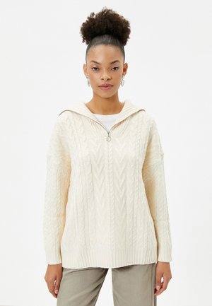 Crème kabelgebreide hoodie met oversized ontwerp, geribde zoom en ritsafsluiting. Heeft een brede kraag en complexe patronen.