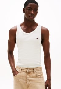 Débardeur en coton blanc avec un col rond, arborant un petit logo sur la poitrine. Assorti à un jean en denim beige clair.