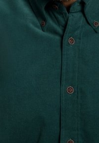 Mörkgrön kordflorsskjorta med knäppning framtill, prydd med bruna knappar och en texturerad tyg. Nära bild av kragen och knappslån.