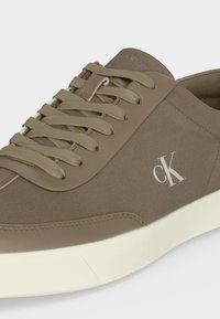 Baskets taupe avec tige en tissu et cuir, lacets beiges, semelle blanche et logo "cK" blanc sur le côté.