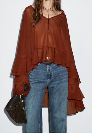 Femme portant un haut ample et transparent couleur rouille avec des manches superposées, jean bleu, tenant un sac à main en daim marron foncé.