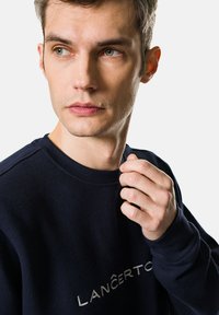 Granatowy sweter z okrągłym dekoltem, z haftowanym logo w jasnoszarym kolorze. Miękka tekstura. Dopasowany krój, długie rękawy.