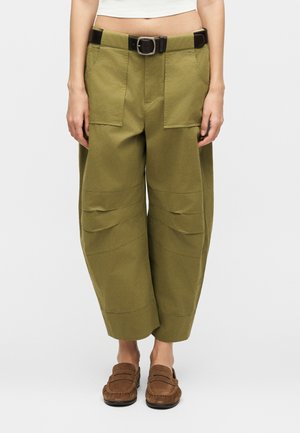 Nő olívazöld, széles szárú, rövidített nadrágot visel nagy első zsebekkel és fekete övvel, barna velúr mokaszinokkal párosítva.