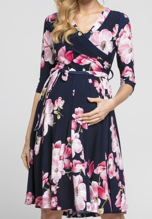 Zwangere vrouw die een marineblauwe wikkeljurk draagt met roze en witte bloemenpatronen, hand zachtjes rustend op haar buik.