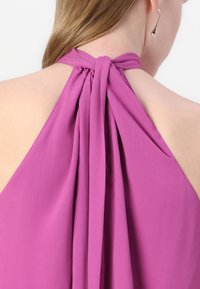 Fialový halterneck top s drapériou a uzlom na dekolte. Hladká textura a otvorený dizajn chrbta bez ozdôb.