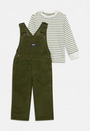 Salopette pour enfant en velours côtelé vert olive avec une poche poitrine, portée sur un t-shirt blanc à manches longues à fines rayures vertes.