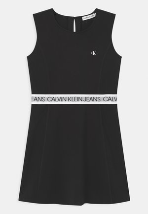 Czarna sukienka bez rękawów z okrągłym dekoltem, białym paskiem w talii z nadrukiem "Calvin Klein Jeans," oraz małym, białym logo CK na piersi.