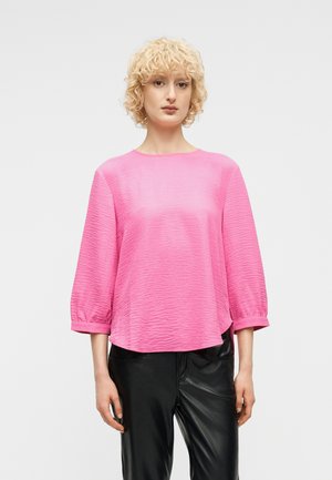 Kobieta o krótkich kręconych blond włosach, ubrana w różowy, długi rękaw o fakturze oraz czarne skórzane spodnie, stojąca na białym tle.