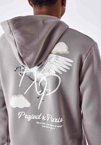 Un sweat à capuche gris avec un grand graphique blanc de pégase au dos, des nuages et du texte indiquant "Project x Paris" dans la partie inférieure.