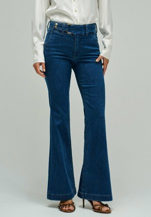 Femme portant un jean évasé bleu foncé taille haute avec des boutons à la taille, associé à un chemisier blanc et des sandales à talons brunes à brides.