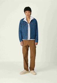 Τζιν μπουφάν με μπλε ξεβάμμα, κούμπωμα με κουμπιά· φορεμένο πάνω από άσπρο πουκάμισο. Καφέ παντελόνι, μπεζ αθλητικά παπούτσια ολοκληρώνουν το ντύσιμο. Απλός, καθημερινός σχεδιασμός.