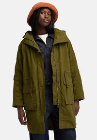 Olivgrön parkajacka med huva, stora framfickor, justerbar dragsko och dragkedjestängning, bärs över ett blått denimplagg.