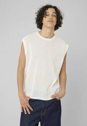 Jeune homme aux cheveux bouclés portant un haut en maille blanc sans manches et un jean bleu foncé, debout avec une main dans la poche.