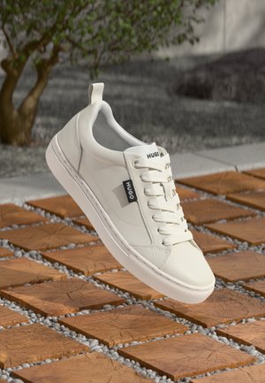 Biały sznurowany sneaker Hugo unoszący się nad drewnianymi kwadratowymi płytkami z małymi szarymi kamykami, plener z drzewem i żwirowym tłem.