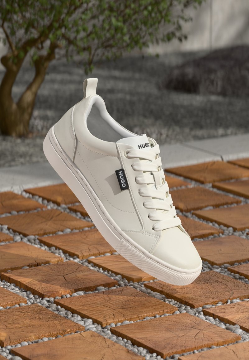 Baltas Hugo sportbačių modelis su raišteliais skrieja virš medinių kvadratinių plytelių su mažais pilkais akmenėliais, lauko aplinka su medžiu ir žvyro fone.