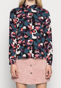 Blusa floreale con bottone su sfondo scuro, caratterizzata da grandi fiori multicolori in rosa, teal e bianco. Maniche lunghe.