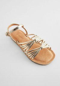 Goldene Riemchensandale mit einer Innensohle aus beige Wildleder, die ein spiralförmiges Akzentdesign und einen verstellbaren Knöchelriemen aufweist. Flache Sohle mit leicht abgerundeter Zehenpartie.