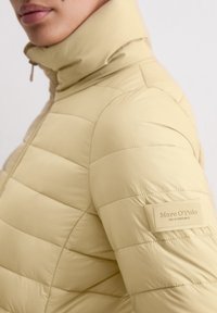 Persoon die een beige gewatteerde jas draagt met een hoge kraag, voorzien van een Marc O'Polo label op de bovenarm.