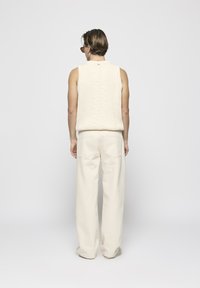 Beige ärmelloser Pullover aus Kabelstrick, kombiniert mit weit geschnittenen beigen Hosen, die eine weiche Textur und einen entspannten Schnitt zeigen.