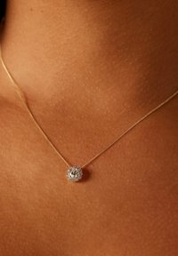 Gouden ketting met een ronde, fonkelende hanger met een centrale steen omringd door kleinere kristallen, afgebeeld tegen een huidskleurige achtergrond.