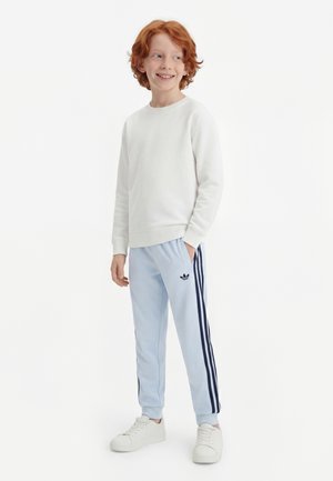 Garçon roux debout, portant un sweat blanc, un pantalon de survêtement Adidas bleu clair avec des rayures bleu marine, et des baskets blanches, regardant sur le côté en souriant.