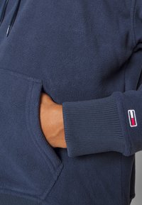 Marineblauwe fleece hoodie met een buidelzak, geribbelde manchetten en een logo-accent. Zachte textuur en een relaxte pasvorm.