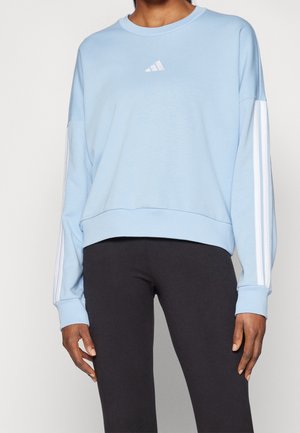 Person trägt ein hellblaues Adidas-Sweatshirt mit weißen Streifen an den Ärmeln und schwarze enge Hosen, steht vor einem einfarbigen Hintergrund.