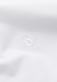 Tissu blanc texturé avec un emblème brodé subtil comportant une couronne de laurier, une couronne et la lettre « O » au centre.