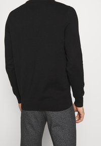 Maglione nero con un motivo a maglia testurizzato, maniche lunghe e scollatura rotonda. Indossato con pantaloni grigi a fantasia, che mostrano un design aderente.