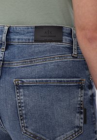 Jeans de mezclilla azul con una etiqueta de cuero negro en la cintura, con costuras contrastantes y dos bolsillos traseros. Textura suave.