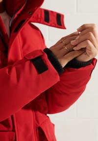 Rode geïsoleerde jas met een ritssluiting aan de voorkant, grote kraag en zwarte ribgebreide manchetten. Bevat zwarte klittenbandsluitingen op de mouwen.