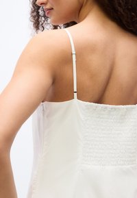 Γυναίκα με σγουρά μαλλιά πο υ φοράει λευκό αμάνικο τοπ με ρυθμιζόμενους ιμάντες και πλεκτή πίσω λεπτομέρεια.