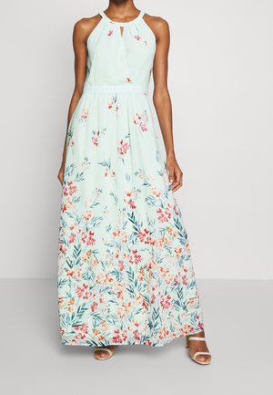 Kobieta ubrana w bezrękawnikową, miętowo-zieloną sukienkę maxi z dekoltem halter i kwiatowym wzorem oraz białe sandały na obcasie na jednolitym tle.