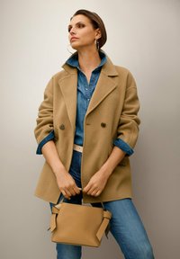 Vrouw in een beige jas en denim shirt met opgerolde mouwen houdt een tan handtas vast, staande tegen een effen lichte muur.